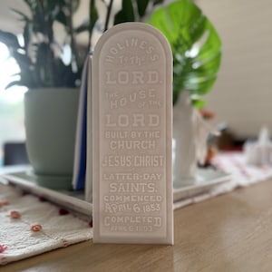 Puede incluir: Una placa rectangular blanca con bordes superiores redondeados. La placa tiene texto en relieve que dice "HOLINESS TO THE LORD" y otro texto religioso. La placa conmemora la construcción de una iglesia, con fechas de inicio y finalización.
