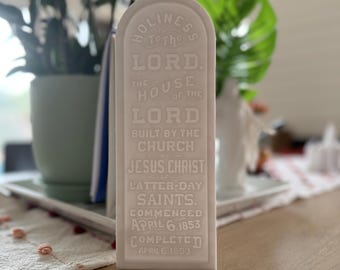 May include: A white rectangular plaque with rounded top edges. The plaque has embossed text that reads "HOLINESS TO THE LORD" and other religious text. The plaque commemorates the construction of a church, with dates of commencement and completion.