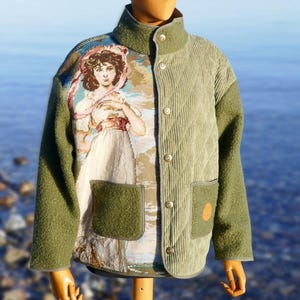 Könnte beinhalten: Olivgrüne Jacke mit einem Wandteppich-Panel, das eine Frau in einem weißen Kleid und einem rosa Hut zeigt. Die Jacke hat eine Cordfront, zwei Taschen und einen hohen Kragen. Die Ärmel und der Rücken sind aus strukturiertem grünem Stoff.