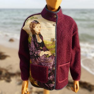 Könnte beinhalten: Burgunderfarbene Jacke mit einem Wandteppich-Panel, das eine Frau in einem lila Kleid zeigt. Die Jacke hat einen hohen Kragen, einen Knopfverschluss und zwei Vordertaschen. Der Besatz ist dunkelblau.