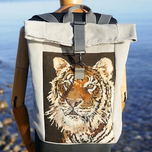 Könnte beinhalten: Beigefarbener Cord-Rucksack mit einem detaillierten, gestickten Tigergesicht auf der Vorderseite. Die Tasche hat einen Rollverschluss, graue Träger und einen schwarzen Verschluss. Der Hintergrund zeigt eine verschwommene Ansicht eines Gewässers.