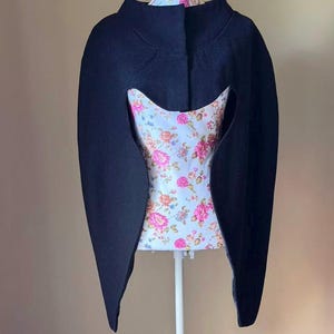 May include: A black cape-style garment with long sleeves and a high collar. The cape has a unique cut-out design in the front, revealing a floral patterned fabric underneath. The floral pattern features pink and white flowers on a light blue background.