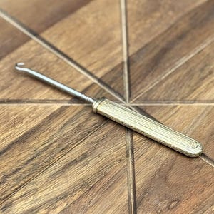 May include: A silver-toned crochet hook with a textured handle lies on a wooden surface. The handle has a detailed, engraved design. The hook is positioned diagonally, with a blurred background of purple flowers.