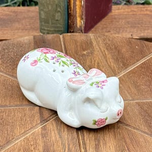 May include: A white ceramic pig-shaped salt and pepper shaker set. The pig is decorated with pink floral patterns and a pink bow. The shaker has two holes on top for dispensing salt and pepper.