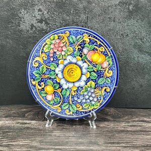 May include: A decorative ceramic plate with a blue rim and a central floral design. The plate features a large white and yellow flower surrounded by colorful fruits and green leaves. The plate is displayed on a clear stand.