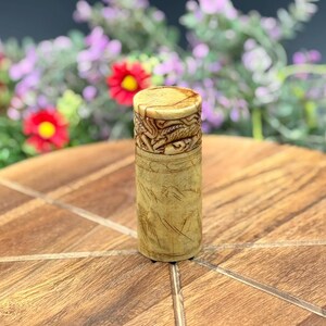 May include: A cylindrical, light-colored wooden container with intricate carvings. The top features detailed relief work, while the body displays a landscape design. The container is standing on a wooden surface, with flowers in the background.