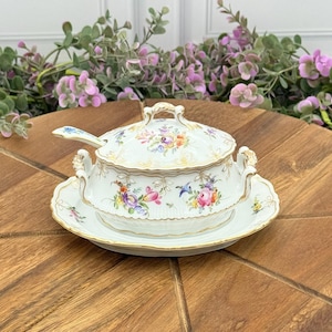 May include: A white porcelain soup tureen with a lid, handles, and a matching saucer. The tureen and saucer are decorated with colorful floral patterns and gold trim. A spoon rests on the tureen. The set is on a wooden surface.