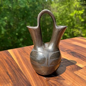 May include: A dark gray ceramic vase with a unique handle and a decorative pattern. The vase has a smooth, polished surface and is displayed on a wooden table. The background is a blurred view of green trees.