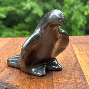 May include: A glossy, black ceramic sculpture of a seal. The seal is depicted in a seated position with its head turned slightly to the side. The sculpture has smooth, rounded features and sits on a wooden surface.