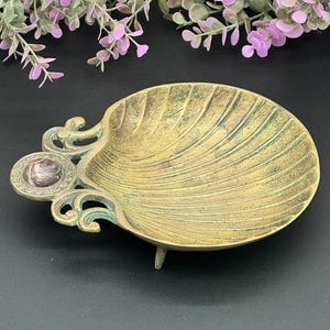 May include: A vintage brass seashell-shaped dish with a decorative edge and a small, round accent. The dish has a weathered, aged appearance with green and gold tones. The dish is supported by three small feet.