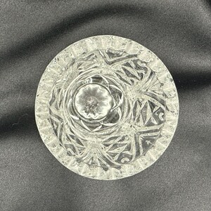 May include: A clear, round ashtray with a detailed, geometric pattern. The ashtray has a central well and a textured rim. The design features repeating shapes and lines, creating a sparkling effect. The ashtray is made of glass.