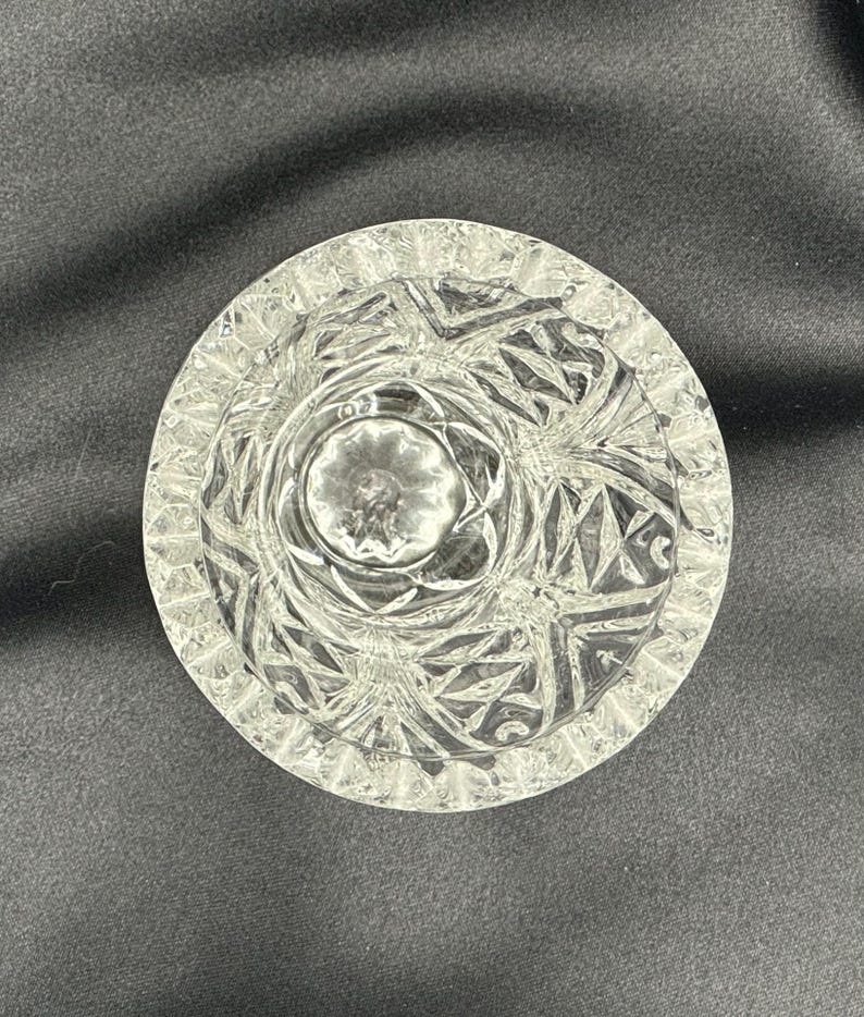 May include: A clear, round ashtray with a detailed, geometric pattern. The ashtray has a central well and a textured rim. The design features repeating shapes and lines, creating a sparkling effect. The ashtray is made of glass.