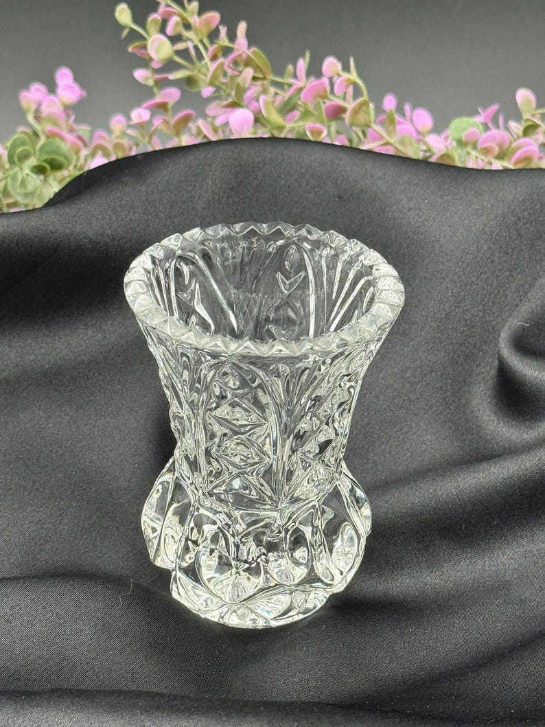 May include: A clear, cut-glass vase with a flared top and a detailed, patterned design. The vase sits on a round base and has a textured surface. The vase is likely used for displaying flowers or other decorative items.