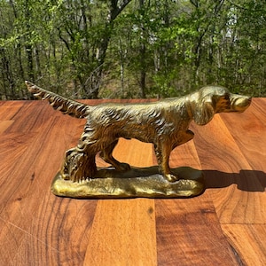 May include: A golden-colored metal sculpture of a dog, likely a setter, standing on a base. The dog has detailed fur and is posed with its tail up. The sculpture is on a wooden surface with a blurred green background.