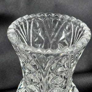 May include: A clear, cut-glass vase with a flared top and a detailed, geometric pattern. The vase has a wide opening and a textured surface, reflecting light. The design features diamond and leaf-like shapes, creating a decorative effect.
