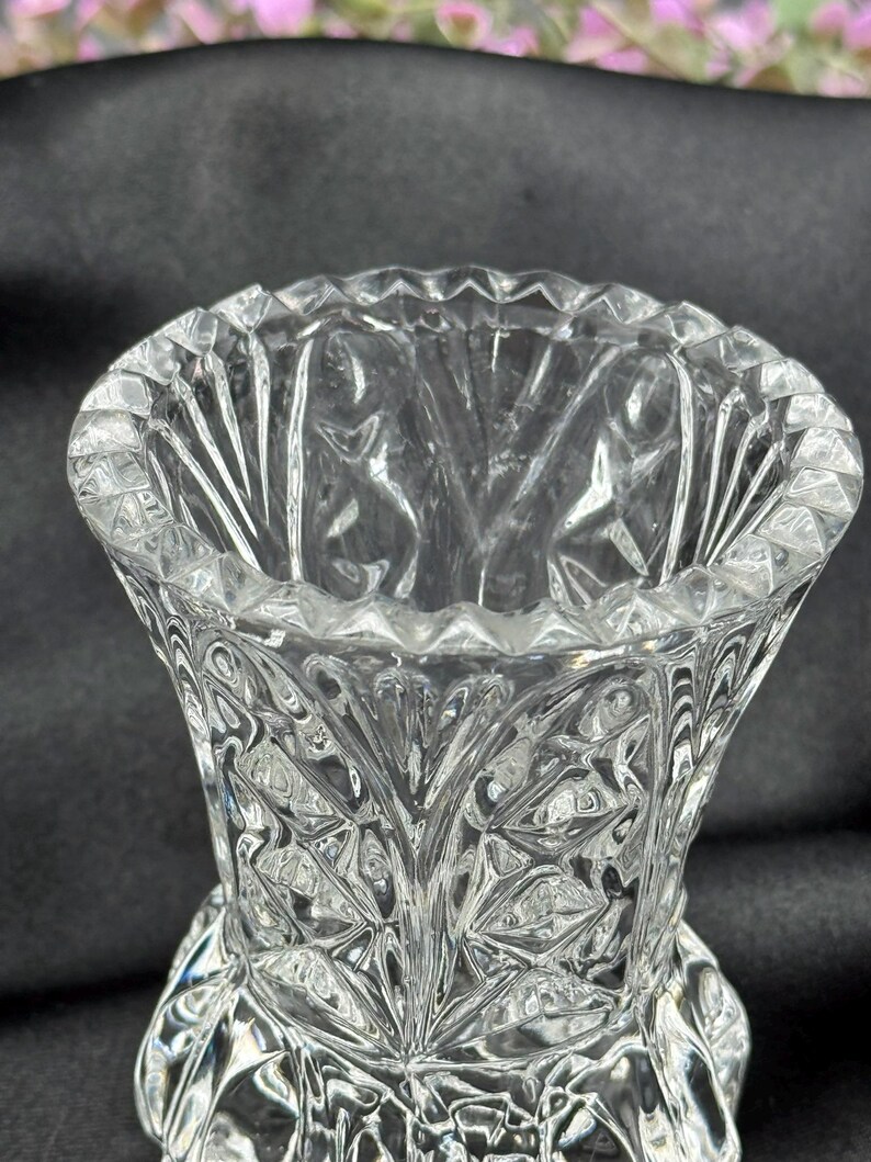 May include: A clear, cut-glass vase with a flared top and a detailed, geometric pattern. The vase has a wide opening and a textured surface, reflecting light. The design features diamond and leaf-like shapes, creating a decorative effect.