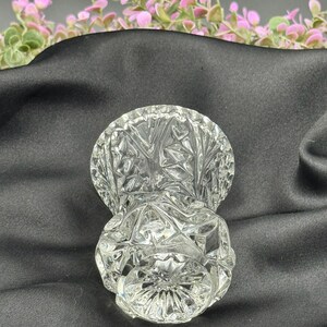 May include: A clear, cut-glass vase with a round base and a flared top. The vase features a detailed, geometric pattern throughout its surface. The background includes pink and green floral accents against a dark fabric.