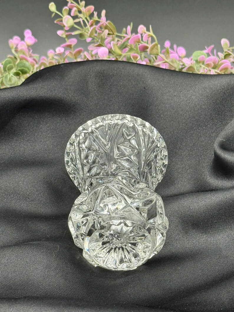 May include: A clear, cut-glass vase with a round base and a flared top. The vase features a detailed, geometric pattern throughout its surface. The background includes pink and green floral accents against a dark fabric.