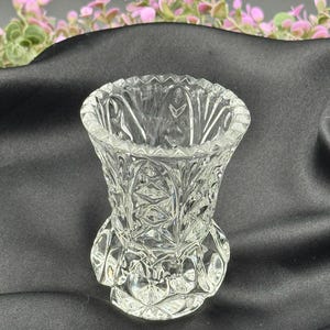 May include: A clear, cut-glass vase with a detailed, geometric pattern. The vase has a flared top and a decorative base. The background is a dark fabric with pink and green floral accents.