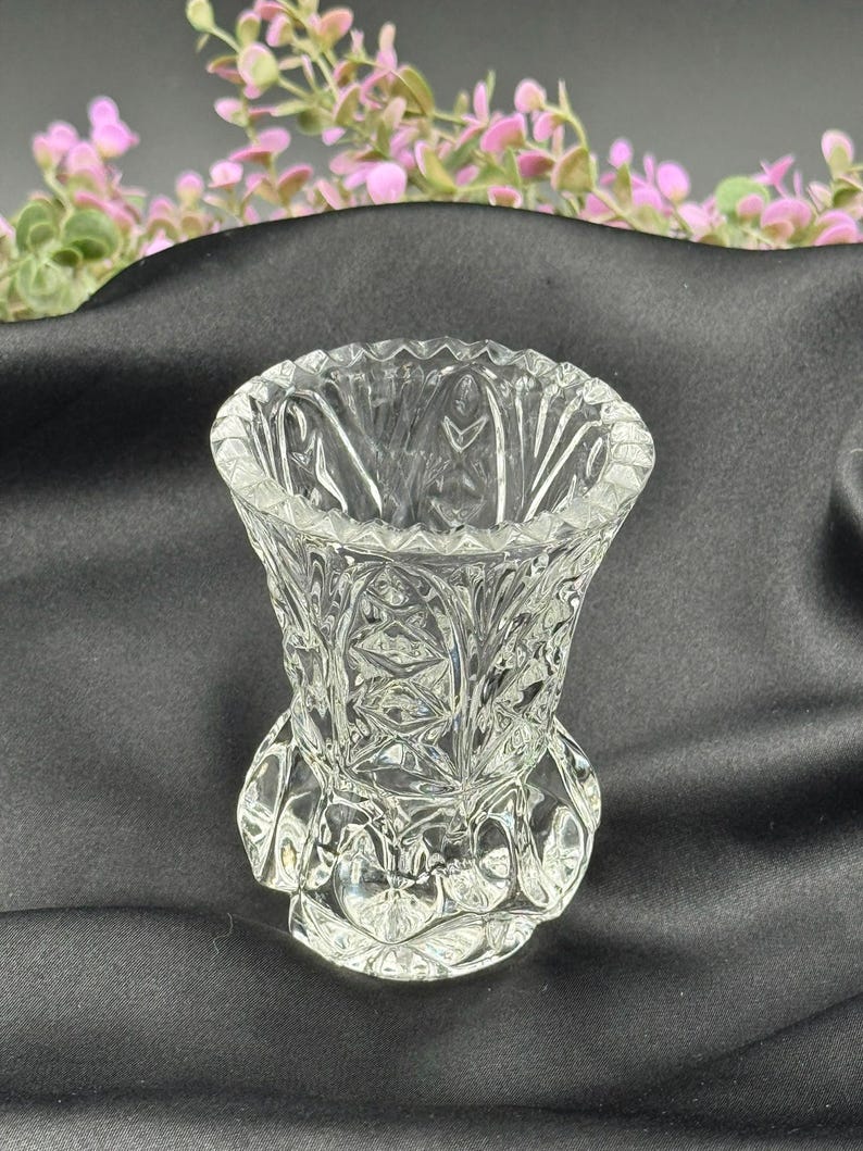 May include: A clear, cut-glass vase with a detailed, geometric pattern. The vase has a flared top and a decorative base. The background is a dark fabric with pink and green floral accents.