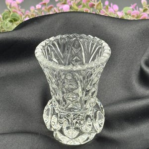 May include: A clear, cut-glass vase with a flared top and a detailed, textured pattern. The vase has a round base and a decorative design around the bottom. The background is a dark fabric with pink and green floral accents.