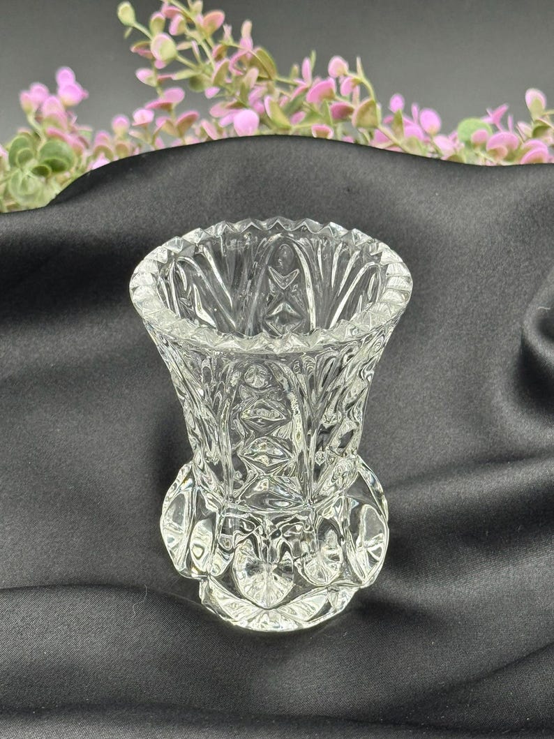 May include: A clear, cut-glass vase with a flared top and a detailed, textured pattern. The vase has a round base and a decorative design around the bottom. The background is a dark fabric with pink and green floral accents.
