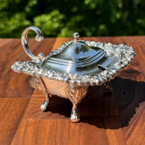 May include: A silver-colored, ornate sauce boat with a lid and handle. The boat has a rectangular shape with decorative floral and scrollwork around the edges. It rests on four small, ornate feet.