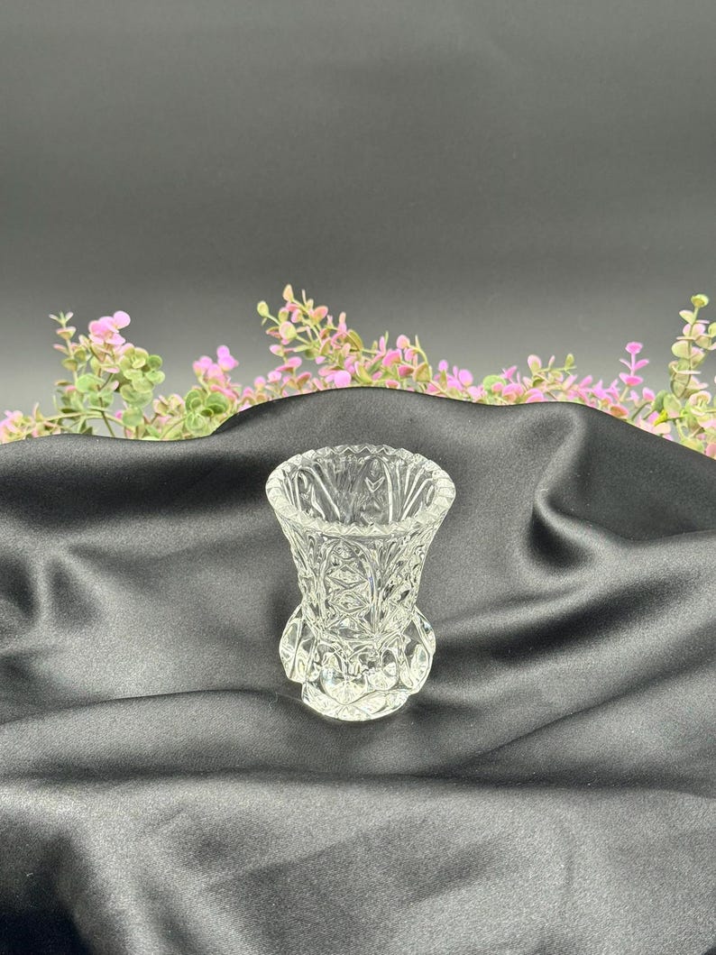 May include: A clear, small, decorative glass vase with a detailed, cut-glass pattern. The vase has a flared top and a rounded base. The background is a black fabric with pink and green floral accents.