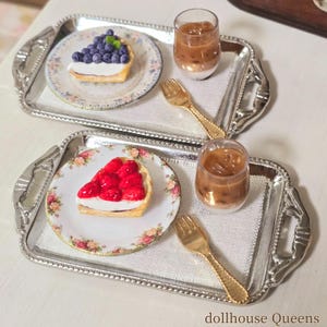 May include: Miniature dessert scene with two silver trays, each holding a fruit tart and a glass of iced coffee. One tart is topped with blueberries, the other with strawberries. Gold forks and floral plates complete the setting.