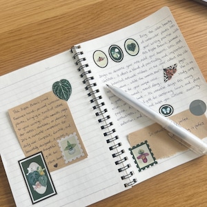 K&ouml;nnte beinhalten: Ein offenes Spiralbuch mit linierten Seiten, das botanische Aufkleber und handschriftlichen Text enth&auml;lt. Ein silberner Stift liegt auf den Seiten. Das Journal enth&auml;lt ein kleines Etikett mit Text und einer botanischen Illustration. Das Thema ist Schreibwaren und Natur.