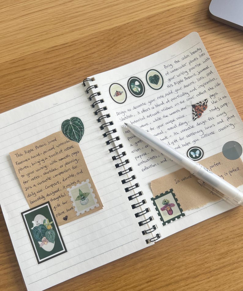 K&ouml;nnte beinhalten: Ein offenes Spiralbuch mit linierten Seiten, das botanische Aufkleber und handschriftlichen Text enth&auml;lt. Ein silberner Stift liegt auf den Seiten. Das Journal enth&auml;lt ein kleines Etikett mit Text und einer botanischen Illustration. Das Thema ist Schreibwaren und Natur.