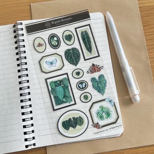 Könnte beinhalten: Ein Spiralnotizbuch mit linierten Seiten, das eine transparente Aufkleberfolie mit botanischen und Schmetterlingsillustrationen zeigt. Ein weißer Stift liegt auf dem Notizbuch. Die Aufkleber zeigen gerahmte Bilder von Blättern und Insekten. Der Text "Ripple Botanic" ist sichtbar.