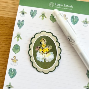 Könnte beinhalten: Ein Notizblock mit grünem Rand und dem Text "Ripple Botanic Watercolor Art and Accessories". Der Notizblock hat einen Aufkleber mit floralem Design und einen weißen Stift. Der Notizblock ist mit botanischen Illustrationen verziert.