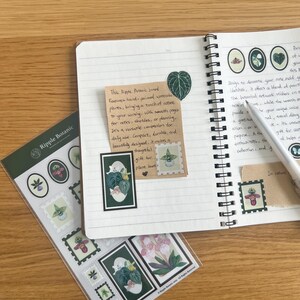 K&ouml;nnte beinhalten: Ein offenes Notizbuch mit botanischen Aufklebern und einer handschriftlichen Notiz. Die Aufkleber mit Pflanzenillustrationen befinden sich in einer transparenten Verpackung. Ein Stift liegt auf den linierten Seiten. Auf einer Holzoberfl&auml;che.