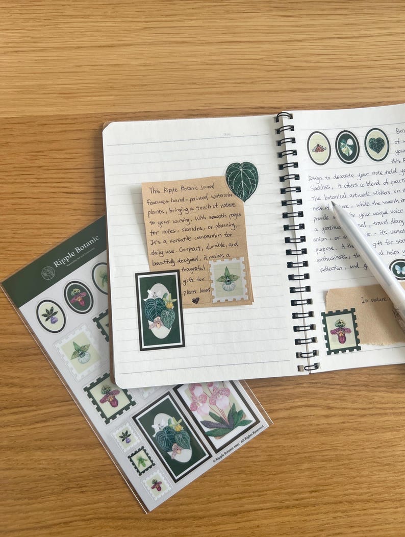 K&ouml;nnte beinhalten: Ein offenes Notizbuch mit botanischen Aufklebern und einer handschriftlichen Notiz. Die Aufkleber mit Pflanzenillustrationen befinden sich in einer transparenten Verpackung. Ein Stift liegt auf den linierten Seiten. Auf einer Holzoberfl&auml;che.