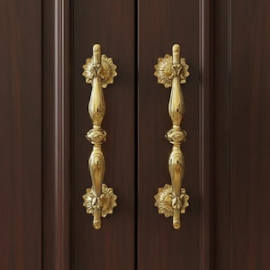 Peut inclure: Deux poignées de placard dorées ornées d'un motif floral. Les poignées sont orientées verticalement et montées sur une porte d'armoire en bois brun foncé. Les poignées ont une esthétique vintage détaillée.