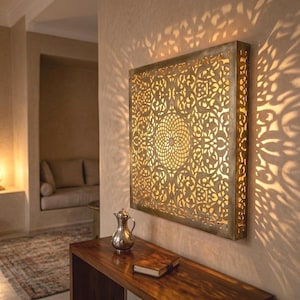 Peut inclure: Une applique murale carrée au fini métallique dotée d'un motif floral complexe découpé au laser. La source lumineuse interne projette un motif d'ombre détaillé et symétrique sur la surface du mur environnant, créant une lueur chaleureuse.