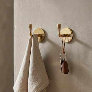 Peut inclure: Deux patères murales en laiton, l'une avec une serviette beige et l'autre avec des clés. Les patères ont une base circulaire et un crochet incurvé. La serviette a un motif brodé. Les clés ont un porte-clés en bois.