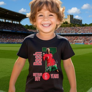May include: A young child wearing a black t-shirt with a graphic of a soccer player in a red jersey. The shirt has the text "Bash Birthday Boy 5 Years" in red and white. The child is smiling in front of a soccer stadium.