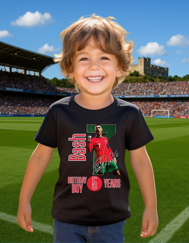 May include: A young child wearing a black t-shirt with a graphic of a soccer player in a red jersey. The shirt has the text "Bash Birthday Boy 5 Years" in red and white. The child is smiling in front of a soccer stadium.