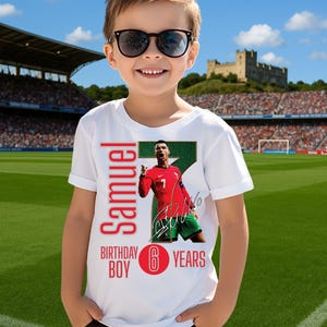 May include: A white t-shirt with a soccer theme. The shirt features a photo of a soccer player in a red jersey, with the name "Samuel", "Birthday Boy", and "6 Years" printed on it. The boy is wearing sunglasses.