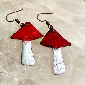 May include: A pair of handmade mushroom earrings. Each earring features a red cap with white accents and a white stem. The earrings have copper-colored ear wires.