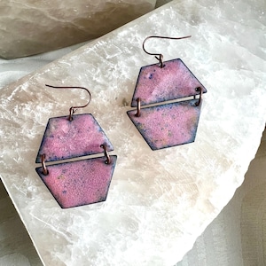 May include: A pair of geometric earrings with a pink and blue color scheme. The earrings are made of metal and feature a two-part design with a copper hook. The earrings are displayed on a white surface.
