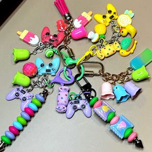 May include: A collection of colorful keychains with various charms. Charms include game controllers, ice pops, candies, and other playful designs. The keychains feature a variety of colors, including pink, blue, green, and yellow, with gold-colored chains and clasps.
