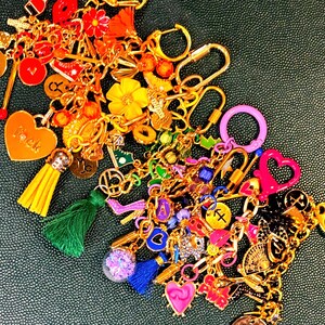 Op de afbeelding: Een verzameling kleurrijke bedels en sleutelhangers in goud, roze, geel, groen en blauw. De bedels omvatten harten, bloemen, kwastjes en andere decoratieve elementen. De achtergrond is een getextureerd donkergroen oppervlak.