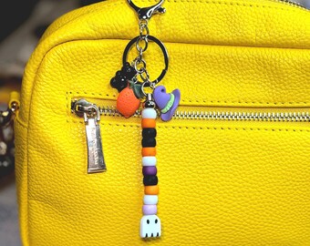 Puede incluir: Un llavero temático de Halloween con un cierre plateado, con dijes de un fantasma, un sombrero de bruja y fruta naranja. El llavero tiene una cadena de cuentas en negro, naranja, blanco, morado y rosa. El fondo es un bolso de cuero amarillo.