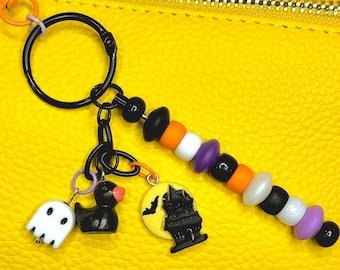 Puede incluir: Un llavero con temática de Halloween con un anillo, cadena y dijes negros. Los dijes incluyen un fantasma blanco, un pato negro con pico rojo y una luna amarilla con una casa embrujada negra. El llavero también tiene una hebra de cuentas negras, naranjas, blancas y moradas.
