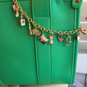 Op de afbeelding: Een felgroene tote bag met een gouden bedelarmband. De armband heeft verschillende bedels, waaronder een hoge hak, parfumflesje en hart. De tas heeft een gestructureerd oppervlak en een handvat.