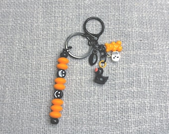 Puede incluir: Un llavero negro con cuentas naranjas y caras sonrientes. El llavero tiene un dije de pato negro, un dije de calavera y un dije de oso amarillo. El llavero está sobre una superficie gris texturizada.
