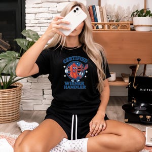 May include: Black t-shirt and shorts set featuring the text "CERTIFIED WIENER HANDLER" and a hot dog graphic with red, white, and blue accents. A person is holding a white smartphone.